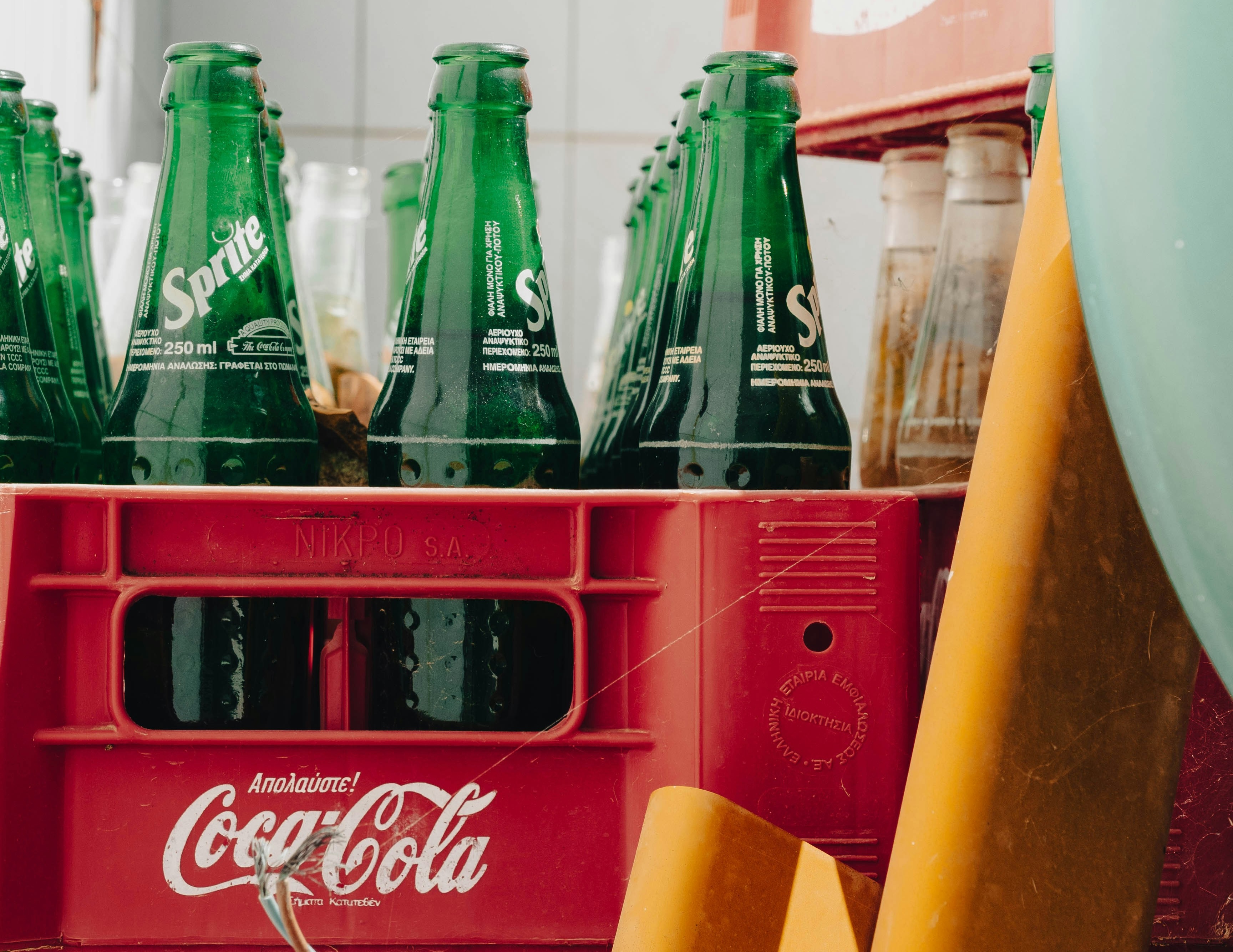 Close-up of green Sprite glass bottles in a red Coca-Cola plastic crate, with a few clear bottles in the background and a large yellow object at the right edge.