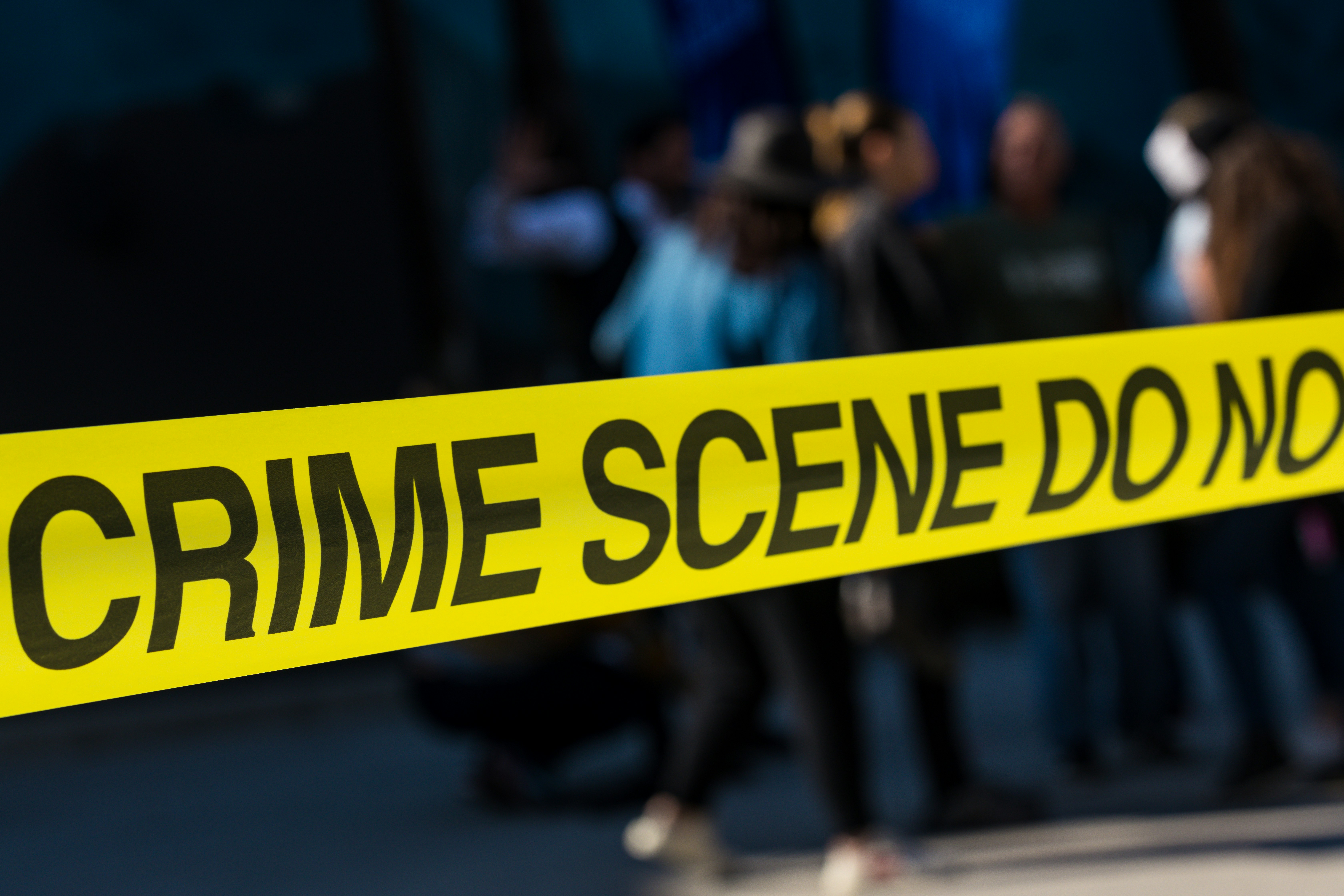 A close-up photograph of yellow crime scene tape in sharp focus reading "CRIME SCENE DO NO[T CROSS]", with a group of blurred figures standing in the background against a dark setting.